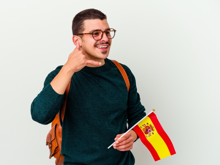 Homme souriant portant un sac à dos, tenant un drapeau espagnol, mimant un geste de conversation téléphonique.