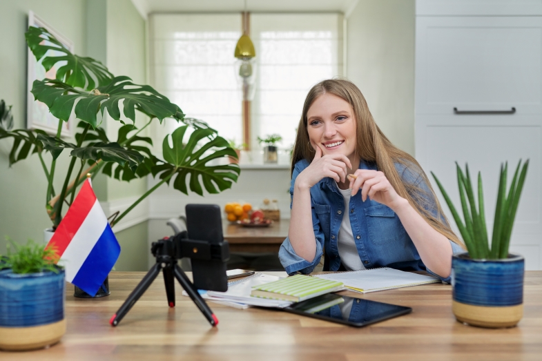 Étudiante assise chez elle, étudiant des cours de néerlandais en ligne et regardant son smartphone avec webcam posé sur trépied